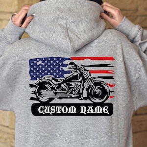 Op de afbeelding: Grijze hoodie met een motorfietsafbeelding en een Amerikaans vlagontwerp. De afbeelding bevat een zwart motorfiets silhouet, rode en witte strepen en een blauw veld met sterren. De tekst "CUSTOM NAME" staat eronder.