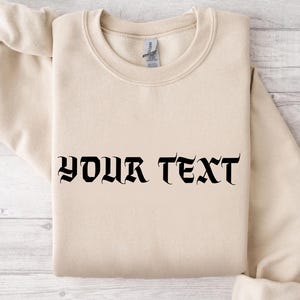 May include: A folded, beige sweatshirt with the words "YOUR TEXT" printed in black gothic lettering across the front. The sweatshirt is made of a soft material and has a crew neck.