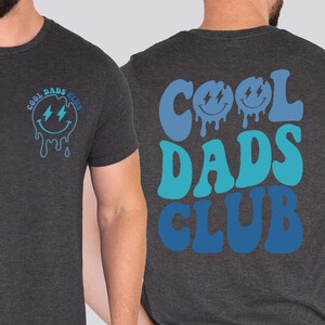 May include: Dark gray t-shirt with "Cool Dads Club" text and a smiling face with lightning bolt eyes. The design is in shades of blue. The same design is on the back of the shirt.