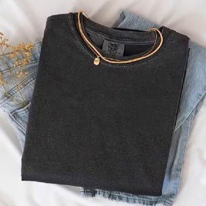 K&ouml;nnte beinhalten: Ein schwarzes T-Shirt mit dem Etikett "CONVRS COLORS", &uuml;ber einer hellblauen Jeans aus Denim. Eine goldene Halskette mit einem kleinen Anh&auml;nger liegt auf dem Shirt. Getrocknete Blumen stecken in der Jeans-Tasche.