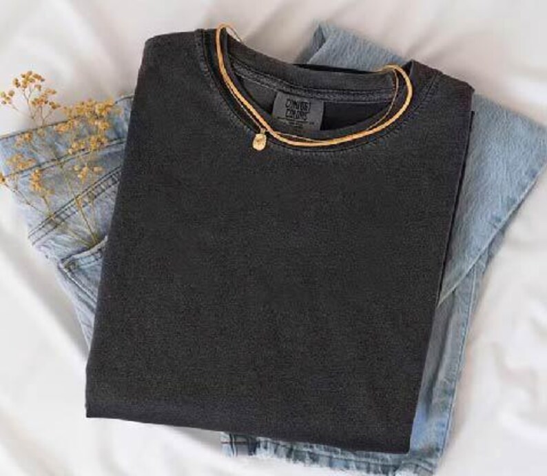 K&ouml;nnte beinhalten: Ein schwarzes T-Shirt mit dem Etikett "CONVRS COLORS", &uuml;ber einer hellblauen Jeans aus Denim. Eine goldene Halskette mit einem kleinen Anh&auml;nger liegt auf dem Shirt. Getrocknete Blumen stecken in der Jeans-Tasche.