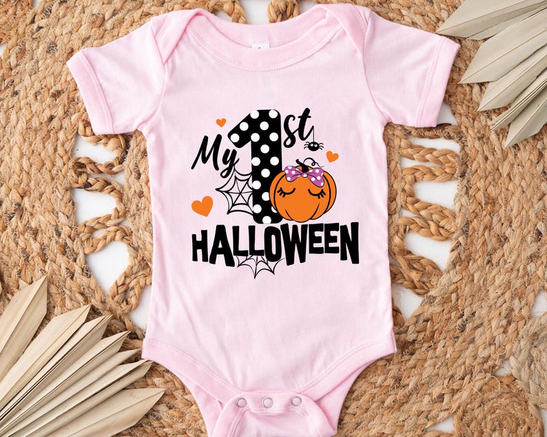 May include: A light pink baby bodysuit featuring a graphic print with the text "My 1st HALLOWEEN". The design includes a black and white polka dot number one, an orange pumpkin with a purple polka dot bow, spider webs, and small orange hearts.