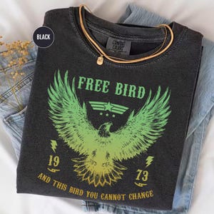May include: Black Comfort Colors t-shirt with a graphic of an eagle and the words "FREE BIRD" in green. The design includes the numbers "19" and "73" and the phrase "AND THIS BIRD YOU CANNOT CHANGE". A gold necklace rests on the shirt.