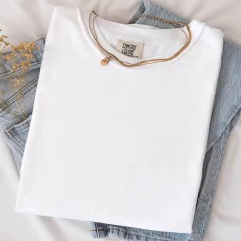 K&ouml;nnte beinhalten: Ein gefaltetes wei&szlig;es T-Shirt mit einem Comfort Colors-Etikett, einer goldenen Halskette und einer blauen Jeans. Das T-Shirt steht im Mittelpunkt, wobei die Jeans und die Halskette einen stilvollen Akzent setzen.