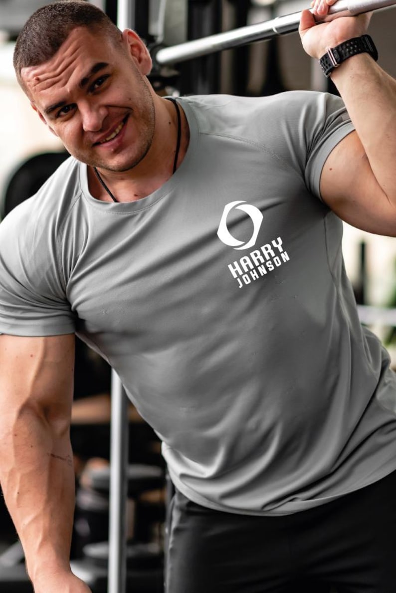 K&ouml;nnte beinhalten: Graues Sport-T-Shirt mit dem wei&szlig;en Text "HARRY JOHNSON". Das Shirt hat einen Rundhalsausschnitt und kurze &Auml;rmel. Das Bild zeigt eine Person, die das Shirt in einem Fitnessstudio tr&auml;gt.