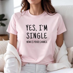 May include: A light pink t-shirt with the text "YES, I'M SINGLE NOW IS YOUR CHANCE" in black. The shirt has short sleeves and a crew neck. The model is wearing a white sweater.