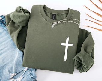 May include: An olive green sweatshirt with a white cross graphic. A silver necklace with a circular pendant rests on the sweatshirt. The sleeves are cuffed. Faded blue jeans are visible in the background.