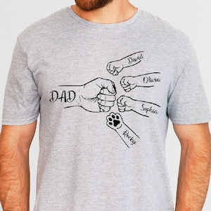 May include: A gray t-shirt featuring a black graphic of a fist bump with the word "DAD" and the names "David", "Olivia", "Sophia", and "Rocky". The design includes a paw print for "Rocky".