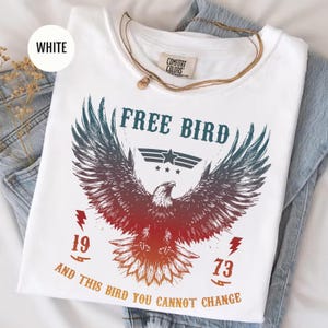 May include: White t-shirt with a graphic of an eagle with outstretched wings, the words "FREE BIRD" above it, and the numbers "1973" below. The shirt also has the text "AND THIS BIRD YOU CANNOT CHANGE". A gold necklace is draped over the shirt.