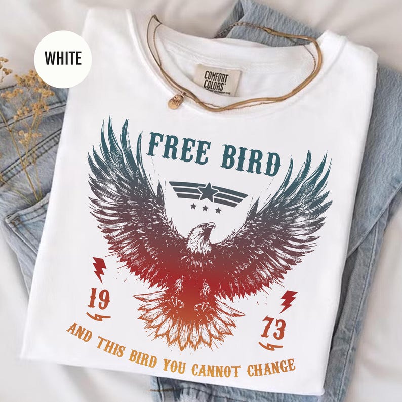 May include: White t-shirt with a graphic of an eagle with outstretched wings, the words "FREE BIRD" above it, and the numbers "1973" below. The shirt also has the text "AND THIS BIRD YOU CANNOT CHANGE". A gold necklace is draped over the shirt.