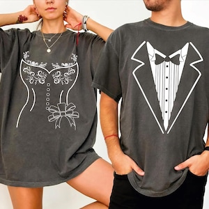 May include: Two charcoal gray t-shirts. One features a white corset design, the other a white tuxedo design. The shirts are oversized and have short sleeves. The designs are simple and graphic, suitable for couples or themed events.