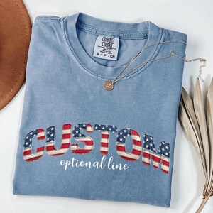 May include: A light blue Comfort Colors t-shirt with the word "CUSTOM" in a red, white, and blue American flag design. The words "optional line" are below the word "CUSTOM". A gold necklace is draped across the shirt.