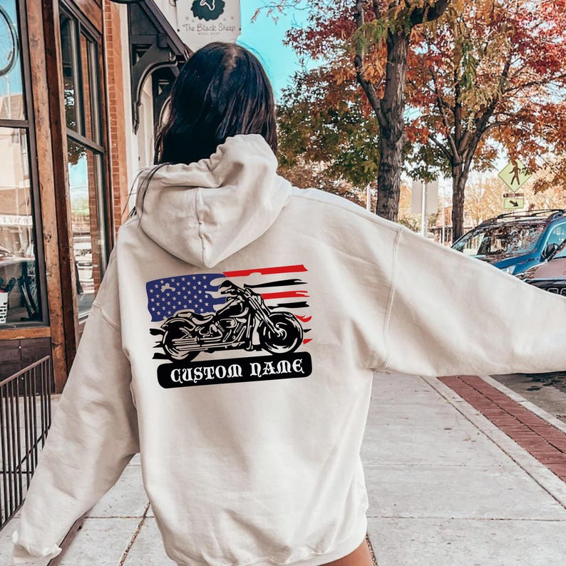 Op de afbeelding: Cr&egrave;mekleurige hoodie met een motorfietsafbeelding en een Amerikaans vlagontwerp. De afbeelding bevat een motorfiets silhouet en de tekst "CUSTOM NAME" in een zwarte banner. De hoodie is een casual kledingstuk.