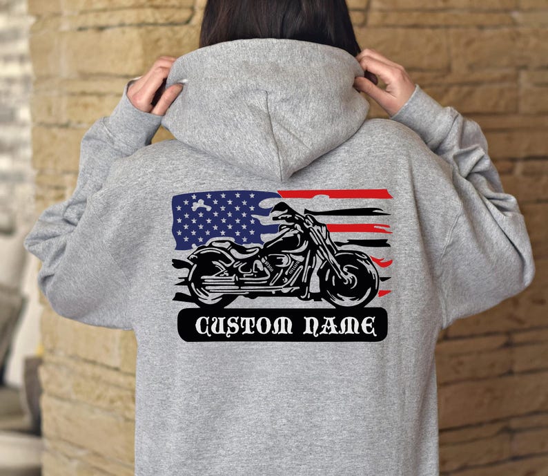 Op de afbeelding: Grijze hoodie met een motorfietsafbeelding en een Amerikaans vlagontwerp. De afbeelding bevat een zwart motorfiets silhouet, rode en witte strepen en een blauw veld met sterren. De tekst "CUSTOM NAME" staat eronder.