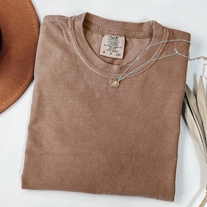 K&ouml;nnte beinhalten: Ein braunes Comfort Colors T-Shirt mit Rundhalsausschnitt, Gr&ouml;&szlig;e S-P-CH. Eine silberne Halskette mit kleinem Anh&auml;nger liegt auf dem T-Shirt. Ein brauner Hut ist teilweise sichtbar. Das T-Shirt ist sauber gefaltet.
