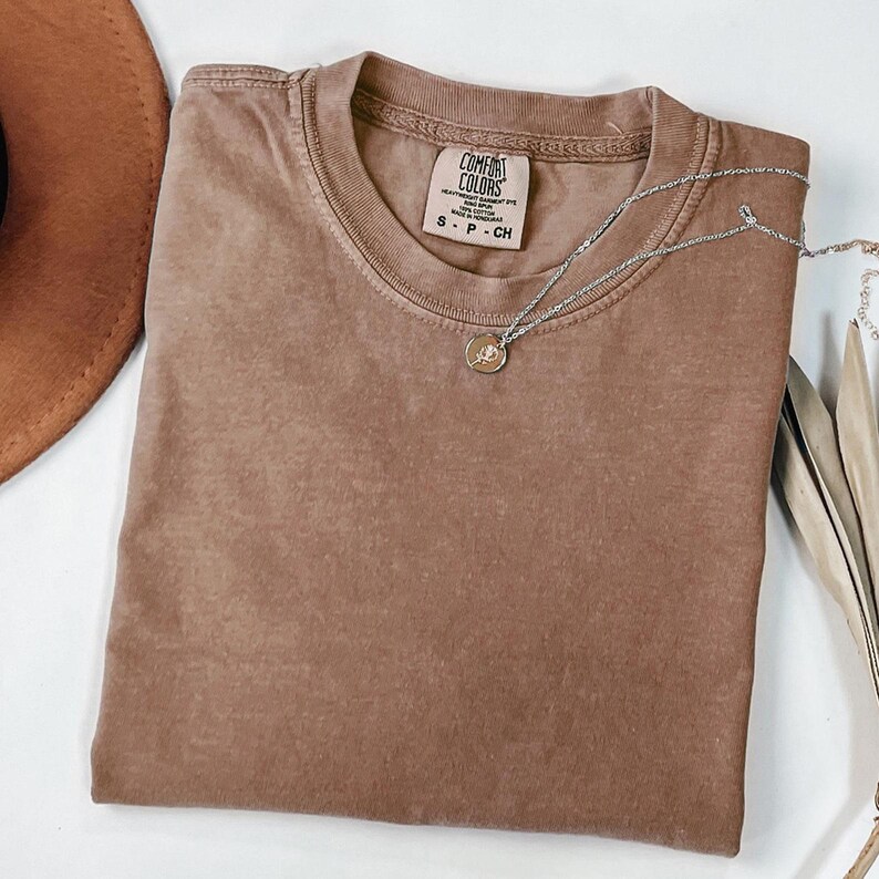 K&ouml;nnte beinhalten: Ein braunes Comfort Colors T-Shirt mit Rundhalsausschnitt, Gr&ouml;&szlig;e S-P-CH. Eine silberne Halskette mit kleinem Anh&auml;nger liegt auf dem T-Shirt. Ein brauner Hut ist teilweise sichtbar. Das T-Shirt ist sauber gefaltet.