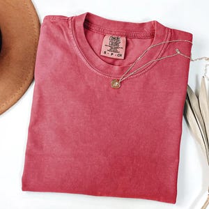 K&ouml;nnte beinhalten: Ein rotes Comfort Colors T-Shirt mit Rundhalsausschnitt, ordentlich gefaltet. Eine goldene Halskette mit Anh&auml;nger liegt auf dem Shirt. Ein brauner Hut und ein getrocknetes Palmblatt sind im Hintergrund. Das Shirt-Etikett lautet "Comfort Colors".