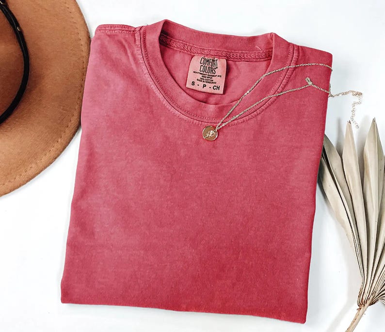 K&ouml;nnte beinhalten: Ein rotes Comfort Colors T-Shirt mit Rundhalsausschnitt, ordentlich gefaltet. Eine goldene Halskette mit Anh&auml;nger liegt auf dem Shirt. Ein brauner Hut und ein getrocknetes Palmblatt sind im Hintergrund. Das Shirt-Etikett lautet "Comfort Colors".