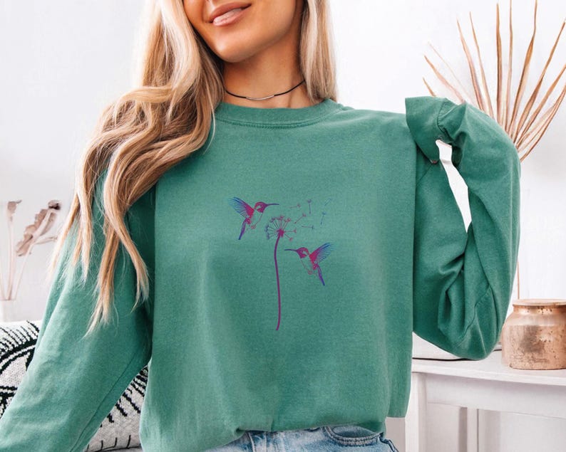 Pu&ograve; includere: Una felpa verde acqua con un disegno di colibr&igrave; e dente di leone. La grafica mostra due colibr&igrave; con ali viola e blu e un becco rosa, che si librano vicino a un dente di leone. La felpa ha maniche lunghe e girocollo.