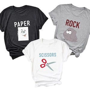 May include: Three t-shirts with Rock, Paper, Scissors designs. The dark gray shirt has "PAPER" and a smiling paper graphic. The gray shirt has "ROCK" and a rock graphic. The white shirt has "SCISSORS" and a scissors graphic.