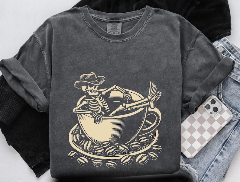 May include: Dark gray t-shirt featuring a skeleton wearing a cowboy hat, relaxing in a coffee cup. The cup sits on a saucer surrounded by coffee beans. The design is in a cream color.