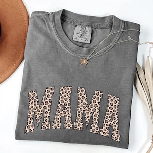 May include: A gray Comfort Colors t-shirt with the word "MAMA" in a leopard print design. A silver necklace with a small pendant is draped across the shirt. A brown hat and dried palm leaves are in the background.