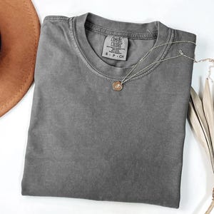 K&ouml;nnte beinhalten: Ein graues Comfort Colors T-Shirt liegt gefaltet auf einer wei&szlig;en Oberfl&auml;che. Eine silberne Halskette mit einem kleinen Anh&auml;nger liegt auf dem Shirt. Ein brauner Hut und getrocknete Palmbl&auml;tter sind im Hintergrund.