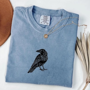 Puede incluir: Una camiseta Comfort Colors azul claro con un dise&ntilde;o de cuervo negro. La camiseta tiene cuello redondo y mangas cortas. Un collar dorado con un colgante est&aacute; drapeado sobre la camiseta. La etiqueta dice "Comfort Colors" y "S-P-CH".