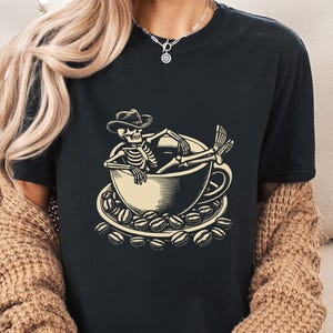 May include: Black t-shirt featuring a beige illustration of a skeleton relaxing in a coffee cup, wearing a cowboy hat. The cup sits on a saucer surrounded by coffee beans. A casual, comfortable tee for coffee lovers.