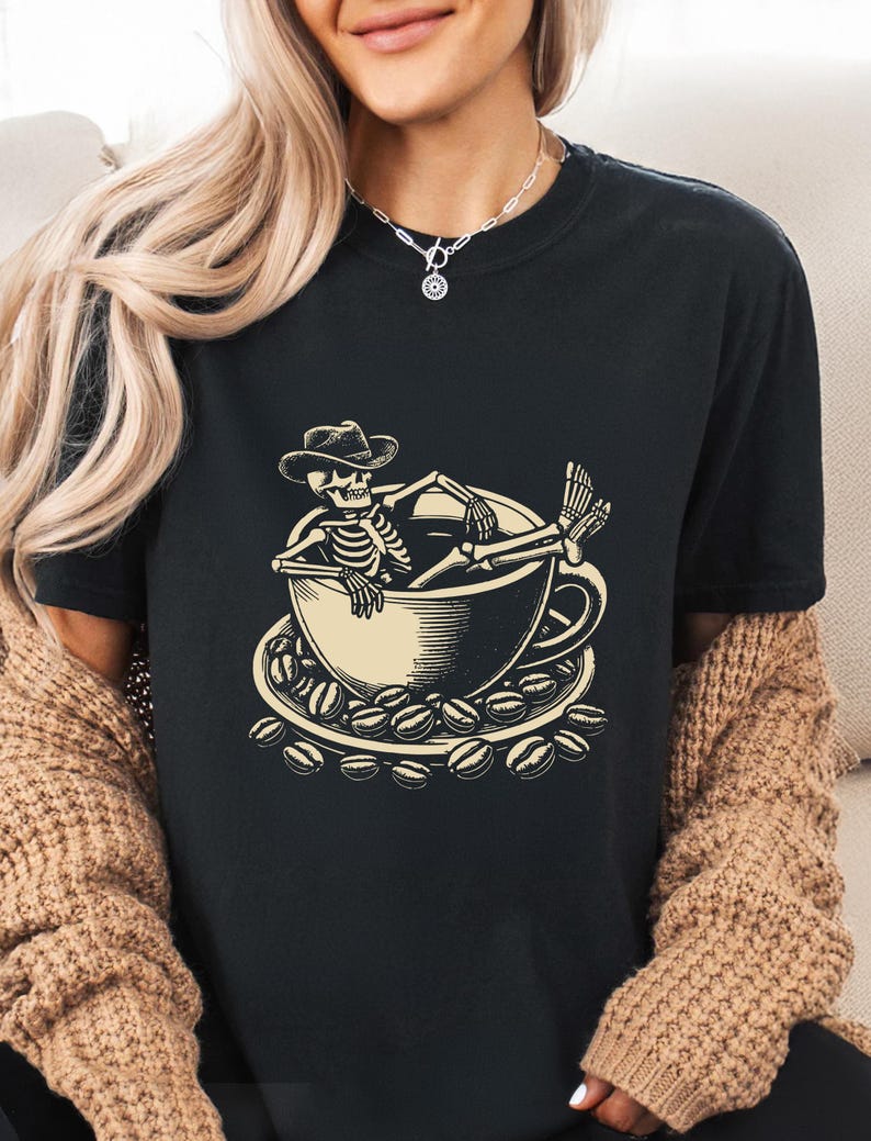 May include: Black t-shirt featuring a beige illustration of a skeleton relaxing in a coffee cup, wearing a cowboy hat. The cup sits on a saucer surrounded by coffee beans. A casual, comfortable tee for coffee lovers.