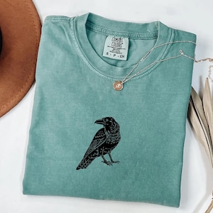 Puede incluir: Una camiseta verde agua con un gr&aacute;fico de cuervo negro. La camiseta tiene cuello redondo y mangas cortas. Un collar plateado con un peque&ntilde;o colgante est&aacute; drapeado sobre la camiseta. La camiseta est&aacute; doblada sobre una superficie blanca.