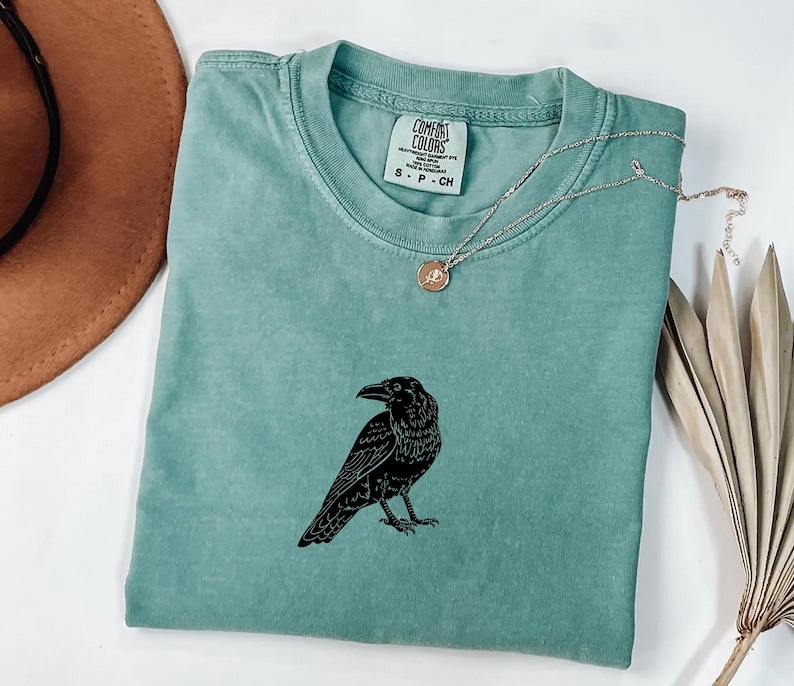 Puede incluir: Una camiseta verde agua con un gr&aacute;fico de cuervo negro. La camiseta tiene cuello redondo y mangas cortas. Un collar plateado con un peque&ntilde;o colgante est&aacute; drapeado sobre la camiseta. La camiseta est&aacute; doblada sobre una superficie blanca.