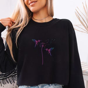 Pu&ograve; includere: Felpa nera a maniche lunghe con un design colorato di colibr&igrave; e dente di leone. L'illustrazione presenta un colibr&igrave; con sfumature viola, blu e rosa e un dente di leone con uno stelo viola.