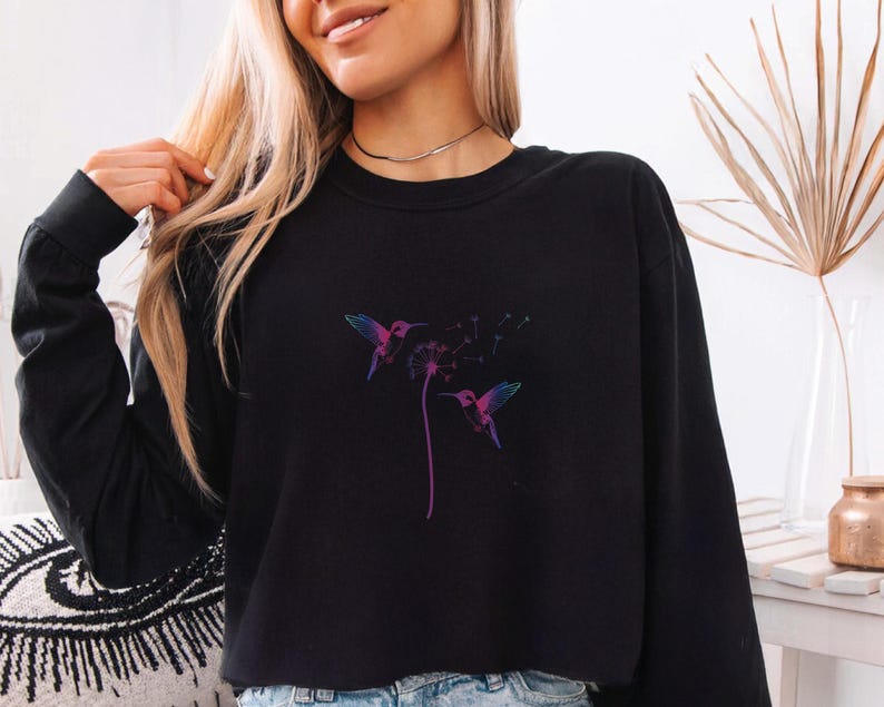 Pu&ograve; includere: Felpa nera a maniche lunghe con un design colorato di colibr&igrave; e dente di leone. L'illustrazione presenta un colibr&igrave; con sfumature viola, blu e rosa e un dente di leone con uno stelo viola.
