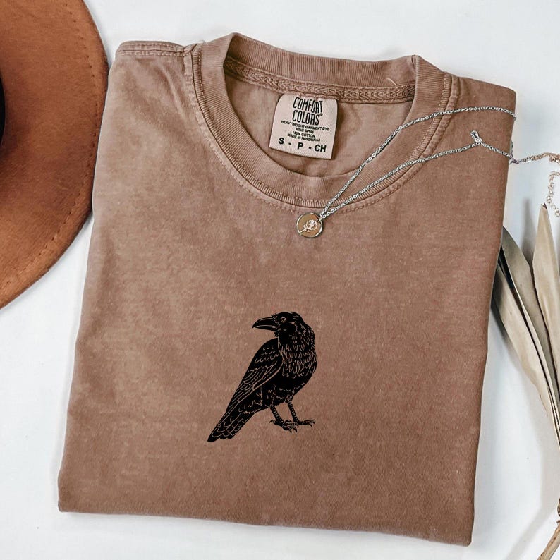 Puede incluir: Una camiseta marr&oacute;n Comfort Colors con un gr&aacute;fico de cuervo negro. La camiseta tiene cuello redondo y mangas cortas. Un collar plateado con un peque&ntilde;o colgante est&aacute; drapeado sobre la camiseta. La camiseta es talla S.
