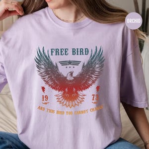 May include: Orchid-colored t-shirt featuring a graphic of an eagle with outstretched wings. The text "FREE BIRD" is above the eagle, with "1973" and the phrase "AND THIS BIRD YOU CANNOT CHANGE" below. The design is in shades of red, orange, and teal.