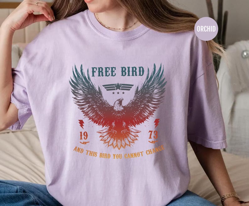 May include: Orchid-colored t-shirt featuring a graphic of an eagle with outstretched wings. The text "FREE BIRD" is above the eagle, with "1973" and the phrase "AND THIS BIRD YOU CANNOT CHANGE" below. The design is in shades of red, orange, and teal.
