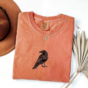 Puede incluir: Camiseta Comfort Colors de color coral con un gr&aacute;fico de cuervo negro. La camiseta tiene cuello redondo y mangas cortas. Un collar dorado con un colgante est&aacute; drapeado sobre la camiseta. Un sombrero marr&oacute;n y una planta seca est&aacute;n en el fondo.