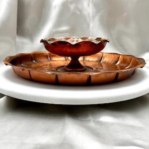May include: A copper serving set featuring a scalloped-edge tray and a small bowl with a pedestal base. The set has a warm, reddish-brown color and sits on a white surface against a white backdrop.