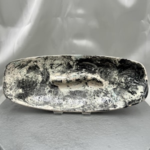 May include: A rectangular ceramic serving dish with a speckled black and white design. The dish has a shallow, elongated shape with a slightly raised center. The surface is textured with a mix of black and white paint, creating a unique, artistic look.