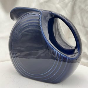 May include: A navy blue ceramic pitcher with a large handle and a curved spout. The pitcher has a glossy finish and a series of concentric ridges around the base. The handle is a large, open loop. The pitcher is a decorative kitchenware item.