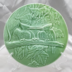 May include: A green ceramic plate with a raised design of two birds perched on a branch. The birds are in a leafy setting with grapes.
