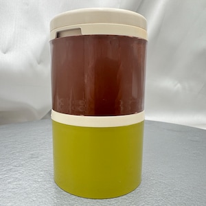 May include: A two-tiered, vintage food container. The top section is a deep brown, the middle section is off-white, and the bottom is a bright green. The lid is off-white. The container is cylindrical and appears to be made of plastic.