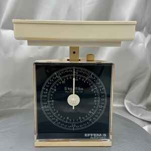 May include: Vintage EFFEM-8 automatic kitchen scale. The scale features a cream-colored tray and a black face with white markings for both kilograms and pounds. The scale can measure up to 5 kg or 11 lbs. The scale has a clear plastic casing.