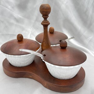 May include: A wooden condiment set with three white ceramic bowls, each with a brown wooden lid and a small wooden knob. The bowls have a woven texture and sit on a wooden tray with a central wooden handle.