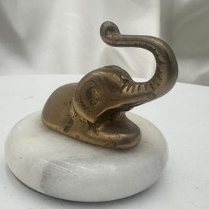 May include: A small, bronze-colored elephant figurine with a curved trunk, resting on a white marble base. The elephant is detailed with visible texture and sits on a round, polished stone.