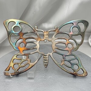 May include: A silver-toned metal butterfly-shaped trivet with openwork wings and body. The trivet has a decorative design and is displayed on a clear stand. The metal has iridescent color variations.