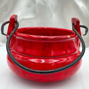 May include: A vibrant red ceramic cauldron with a glossy finish. It features two curved black metal handles attached to the sides. The cauldron has a rounded shape and is likely used for decorative purposes or as a container.
