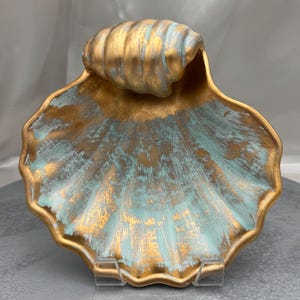 May include: A decorative bowl shaped like a large seashell, featuring a textured finish with a blend of metallic gold and soft teal paint. The interior surface displays a radial pattern of ridges, while the top edge has a prominent, rounded shell detail.