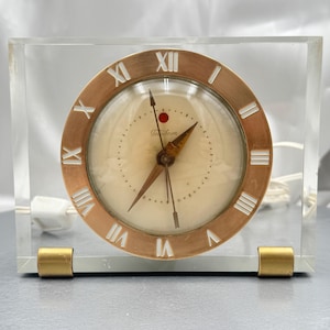 May include: A vintage Techron clock encased in a clear acrylic block. The clock face is cream-colored with Roman numerals and gold-tone hands. The clock has a gold-tone bezel and base supports. A white power cord extends from the back.